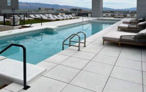 A pool surrounded by a white tiled patio, numerous lounge chairs in grass areas, and an outlook of a city with mountain tops.