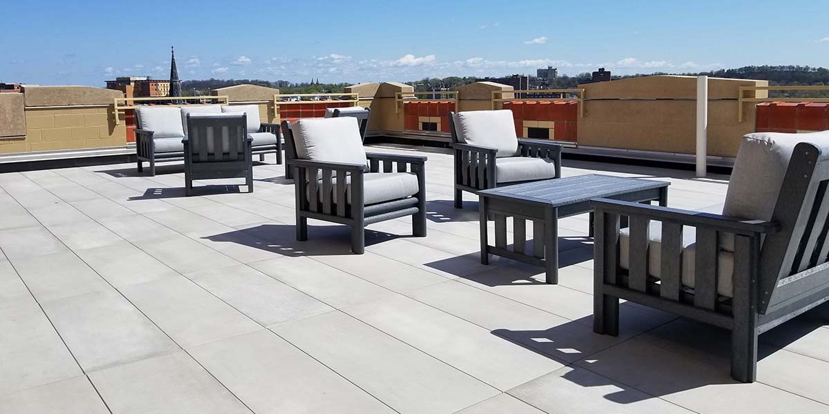 A spacious rooftop patio with porcelain plank decks features several cushioned armchairs and rectangular coffee tables. The area is surrounded by short walls, offering a stunning view of the distant cityscape under a clear blue sky.