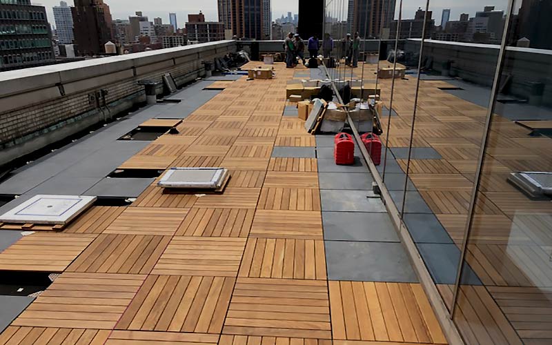 A rooftop being surfaced with Ipe wood tiles, surrounded by a tall railing, featuring construction workers and a skyline view.