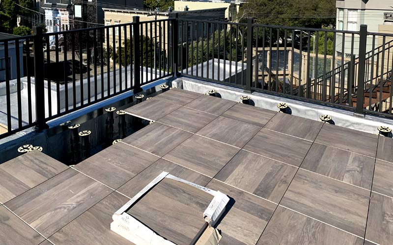 A rooftop patio is under construction, featuring Nut wood look tiles being expertly installed on adjustable pedestals. Black metal railing elegantly surrounds the area, enhancing safety while framing a stunning cityscape. It's an inspiring project for anyone looking to build a rooftop deck.