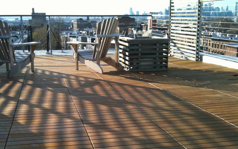 A rooftop deck surfaced with Ipe wood tiles placed in a horizontal pattern, featuring a pair of outdoor chairs facing a skyline.