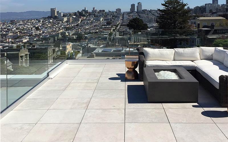 A rooftop deck surfaced with porcelain pavers has a set of furniture, and a fire pit surrounded by a glass railing and a view of the city.