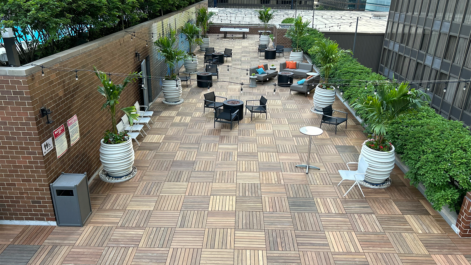 ipe structural deck tiles on a large roof terrace with a brick wall on left, foliage adn bushes on the right and chairs and couches on the deck surface