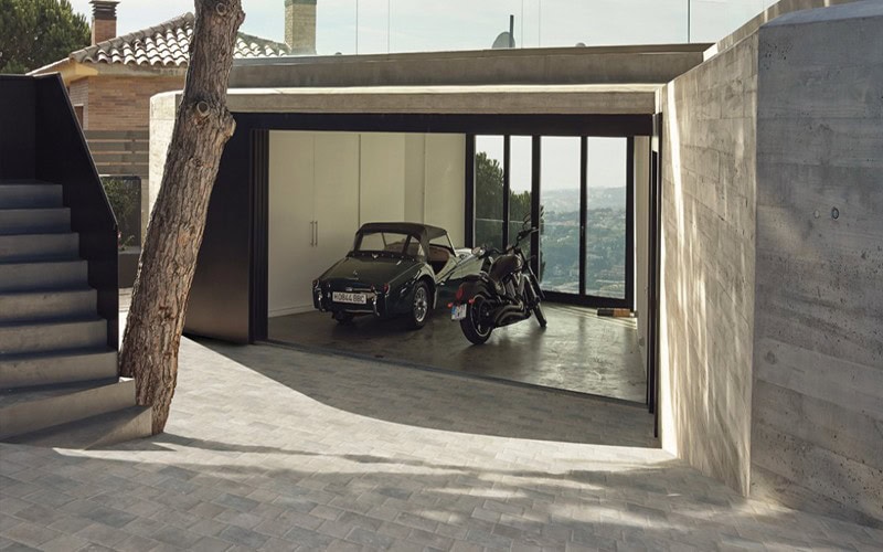 An open modern garage featuring a car and motorcycle looking out to a skyline, surrounded by a stone building.