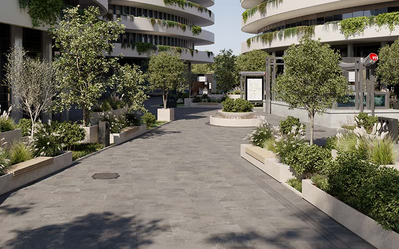 A porcelain paved sidewalk surrounded by cream-colored plant troughs, greenery, tall cream-colored buildings, and signs.