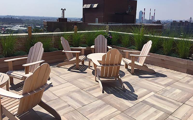 A rooftop deck features outdoor furniture surrounded by troughs of plants and view of a city.