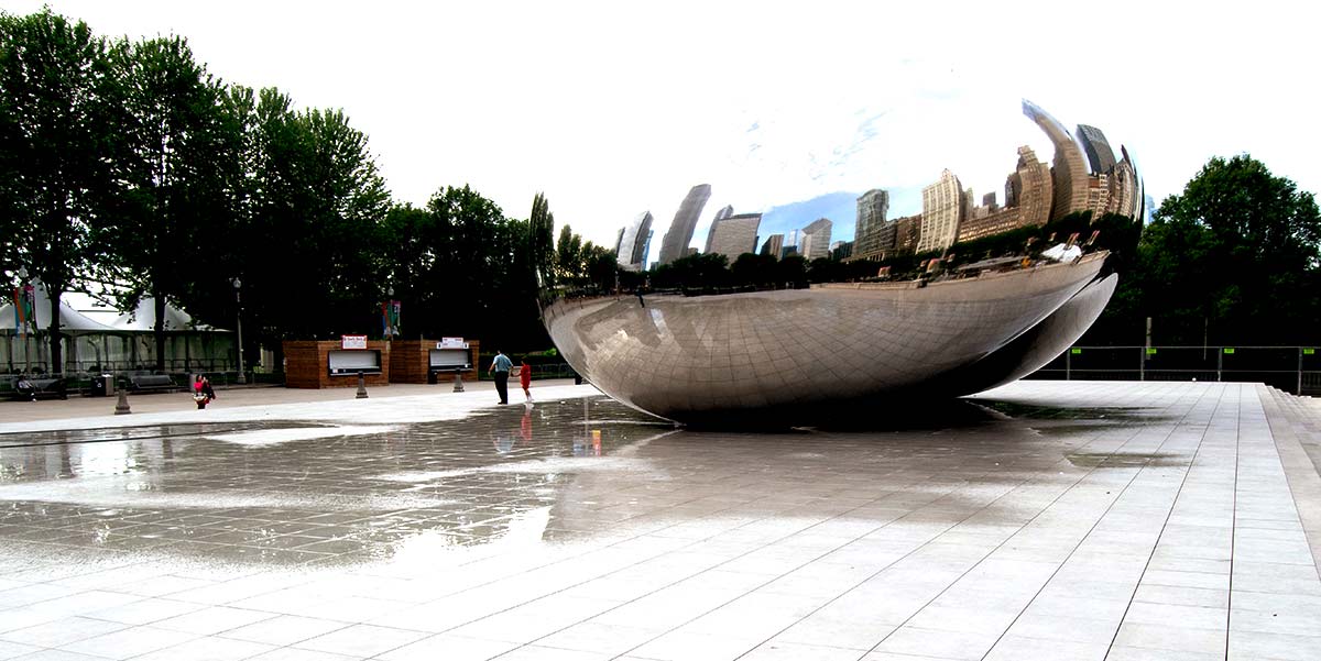 A large, reflective, bean-shaped sculpture sits in an open plaza, mirroring the city skyline and a few people strolling nearby—much like the thoughtful considerations when beginning a porcelain paver installation. Trees and buildings rise in the background under a cloudy sky.