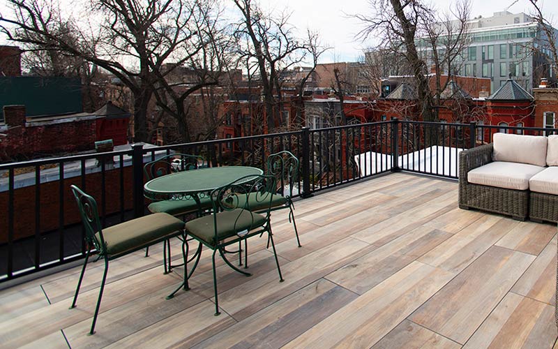 A deck featuring green outdoor furniture surrounded by a black railing and view of bare trees and the tops of houses.