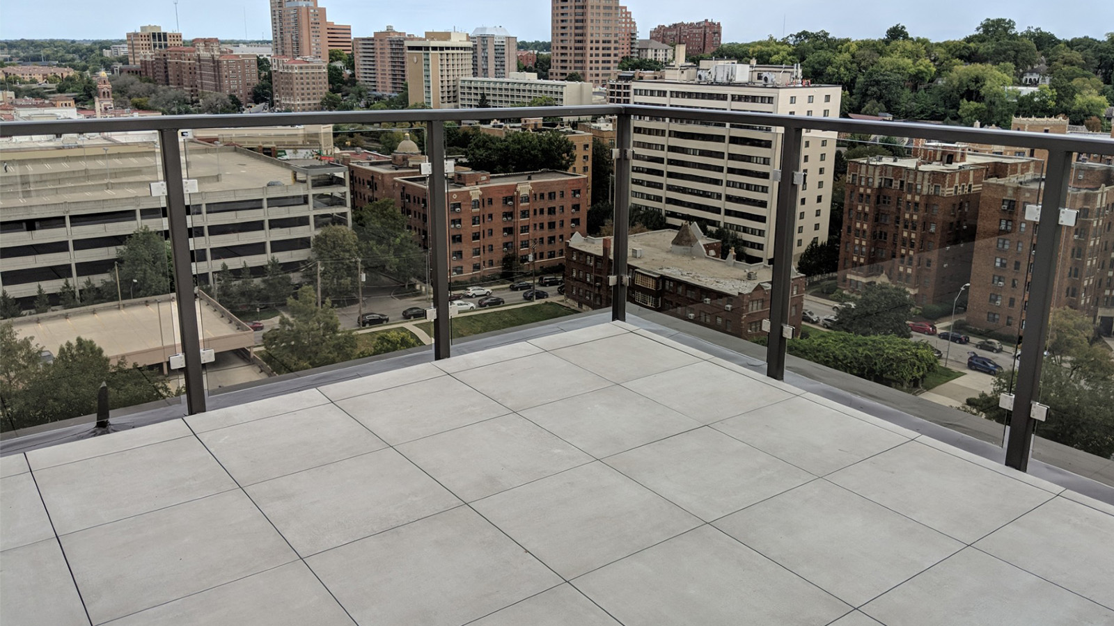 a balcony with gray porcelain pavers and clear glass railing overlooking urban landscape with buildings.