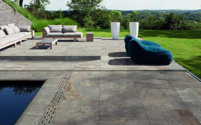 Side view of a modern stone patio, located in a backyard surrounded by grass, featuring outdoor furniture and a pool.